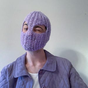 Puede incluir: Una persona lleva una balaclava de punto morado con agujeros para los ojos y la boca. La persona también lleva una chaqueta acolchada morada.