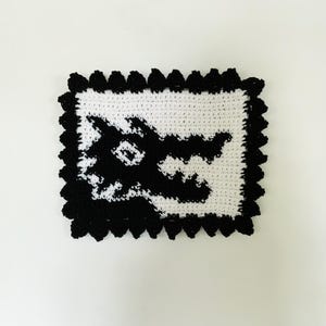 May include: Black and white crocheted square with a wolf face design. The wolf's face is outlined in black and the background is white.