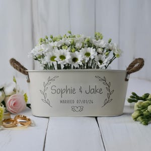 May include: Cream metal planter with rope handles, filled with white flowers. The planter is inscribed with "Sophie & Jake" and the wedding date. A decorative floral arrangement for a wedding or anniversary.