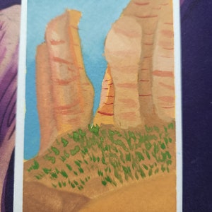 May include: Watercolor painting of a desert landscape featuring tall, reddish-brown rock formations against a light blue sky. Green shrubbery is at the base of the rocks, and the foreground is a light brown color. The painting is on a white rectangular card.
