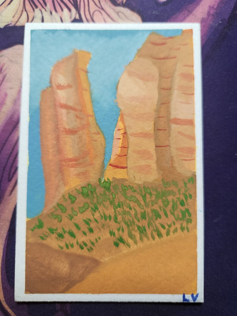 May include: Watercolor painting of a desert landscape featuring tall, reddish-brown rock formations against a light blue sky. Green shrubbery is at the base of the rocks, and the foreground is a light brown color. The painting is on a white rectangular card.