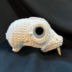 Puede incluir: Una calavera de ganchillo de color blanquecino con una gran abertura circular y un solo diente puntiagudo. La calavera está hecha de hilo texturizado, y el diseño es probablemente hecho a mano.