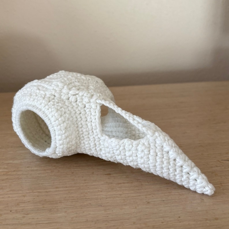 May include: White crocheted bird skull with a hole for the eyes. The skull is made of white yarn and has a long beak.