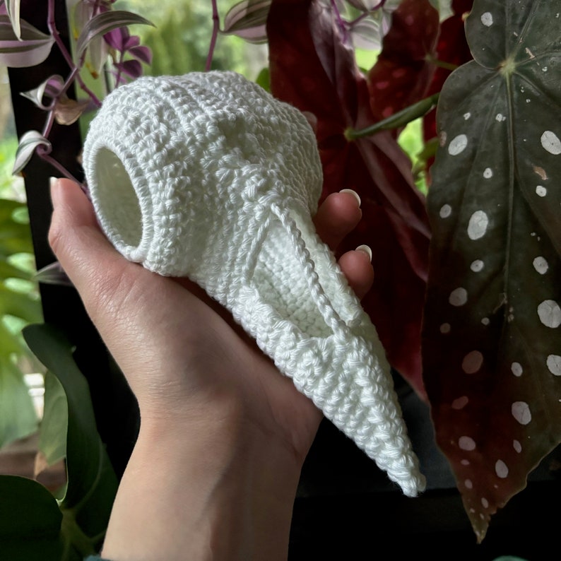 May include: A white crocheted bird skull with a hollowed-out eye socket. The skull is held in a hand against a background of green and red foliage.