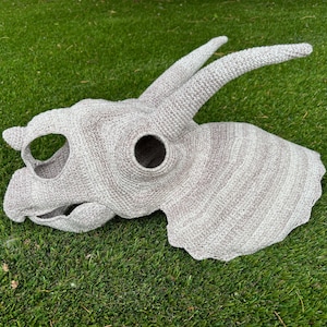 May include: A crocheted grey and white dinosaur head with a large horn. The dinosaur head is made of yarn and has a textured surface.