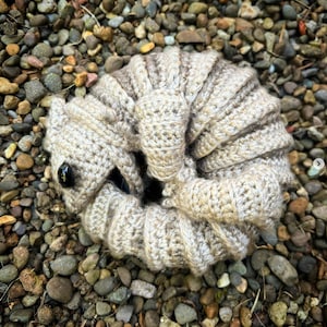 Op de afbeelding: Een gehaakt amigurumi-speelgoed in de vorm van een opgerold wezen met een bruin oog. Het speelgoed is gemaakt van beige garen en heeft een textuur.