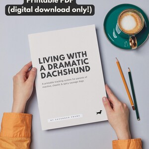 Può includere: Una persona tiene in mano un libro intitolato "Living With A Dramatic Dachshund". Il libro presenta un disegno di un bassotto e il testo "Printable PDF (digital download only!)". Sono visibili anche una tazza di caffè e delle matite.