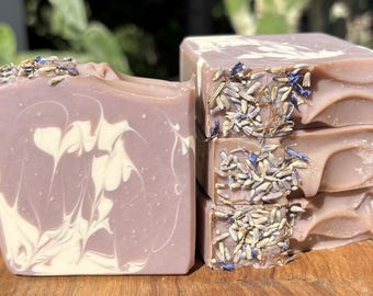 May include: Close-up of handmade lavender soap bars. The soap has a light purple hue with white swirls and is topped with dried lavender buds. The bars are stacked on a wooden surface.
