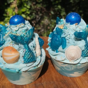 May include: Two blue and white bath bombs shaped like cupcakes with mermaid tails, seashells, and a blue turtle on top.