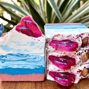 May include: Two bars of handmade soap with a beach theme. The soap is layered with pink, white, and blue swirls. The top layer of the soap has a pink seashell design. The soap is decorated with small seashells.