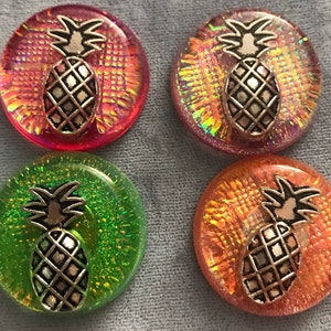 May include: Four round, iridescent, decorative buttons with a pineapple design. The buttons are in shades of pink, orange, green, and yellow.