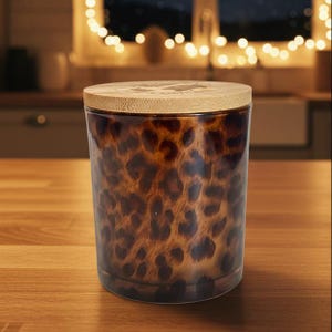 May include: A cylindrical glass jar with a leopard print design in shades of brown and tan. The jar has a wooden lid. The jar is sitting on a wooden surface.
