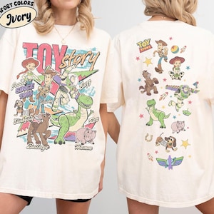 May include: Ivory-colored t-shirt featuring a colorful graphic of Toy Story characters. The front design includes the words "Toy Story" and various characters. The back of the shirt has additional character illustrations. The shirt has short sleeves.