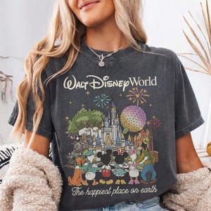 Puede incluir: Camiseta gris oscuro con un gr&aacute;fico colorido de Walt Disney World con el texto "Walt Disney World" y "The happiest place on earth". El gr&aacute;fico incluye personajes de dibujos animados, un castillo y fuegos artificiales.