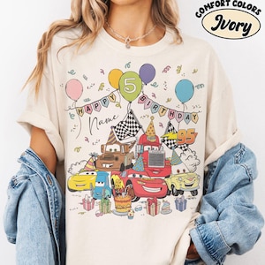 K&ouml;nnte beinhalten: Elfenbeinfarbenes T-Shirt mit einem farbenfrohen Cartoon-Design von Cars-Charakteren, die einen Geburtstag feiern. Das Design umfasst Luftballons, eine Geburtstagstorte und den Text "HAPPY BIRTHDAY" und "5". Das Shirt ist aus Comfort Colors Material.
