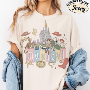 Puede incluir: Camiseta de color marfil con un gr&aacute;fico colorido que presenta personajes de dibujos animados, un castillo y la palabra "m&aacute;gico". La camiseta tiene un corte holgado y mangas cortas. El texto "Comfort Colors" e "Ivory" est&aacute; impreso en la parte superior.
