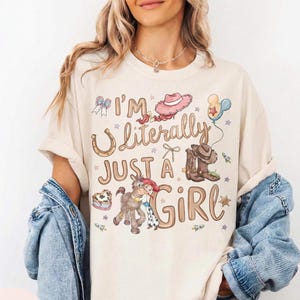 K&ouml;nnte beinhalten: Elfenbeinfarbenes T-Shirt mit dem Text "I'm Literally Just a Girl" und Cartoon-Grafiken eines Hufeisens, Cowboy-Stiefeln, einem Pferd und einem rosa Cowboyhut. Das Shirt hat eine lockere Passform.