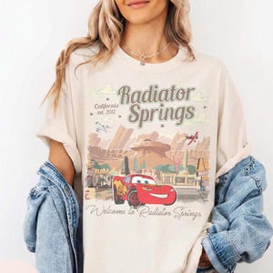 May include: Ivory t-shirt with a graphic of Radiator Springs, California, featuring the text "Radiator Springs" and "Welcome to Radiator Springs." The design includes a red car and a desert landscape.