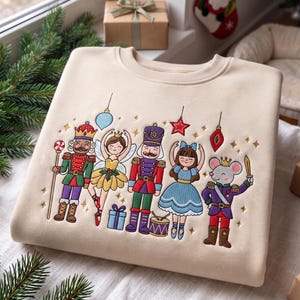 Könnte beinhalten: Ein beigefarbenes Sweatshirt mit Rundhalsausschnitt und einer bunten Stickerei von Nussknacker-Figuren, darunter ein Soldat, eine Fee, ein Trommler, ein Mädchen und eine Maus, verziert mit hängenden Ornamenten, kleinen Sternen und Geschenken.