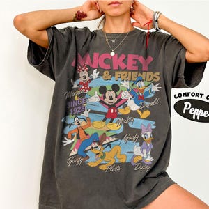 Peut inclure: T-shirt gris fonc&eacute; avec Mickey Mouse et ses amis. Le graphisme comprend Mickey, Minnie, Donald Duck, Dingo, Pluto et Daisy Duck. Le texte comprend "Mickey & Friends" et "Since 1928". Le t-shirt est de Comfort Colors.