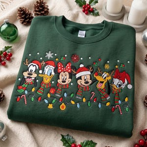 Puede incluir: Una sudadera de cuello redondo en color verde oscuro con un gráfico bordado de personajes de Disney, incluyendo a Goofy, el Pato Donald, Minnie Mouse, Mickey Mouse, Pluto y Daisy, usando bufandas y gorros de invierno, con luces navideñas.