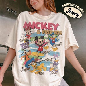 Peut inclure: T-shirt ivoire avec un motif de Mickey Mouse et ses amis. Le design comprend Minnie Mouse, Donald Duck, Dingo, Pluto et Daisy Duck. Le texte "Mickey & Friends Since 1928" est &eacute;galement visible.