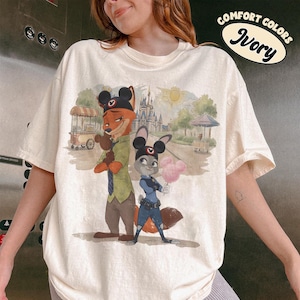 Puede incluir: Camiseta de color marfil con una ilustraci&oacute;n de dibujos animados de Nick Wilde y Judy Hopps de Zootopia, con orejas de Mickey Mouse. El gr&aacute;fico incluye un castillo, un sol y el texto "Comfort Colors Ivory".
