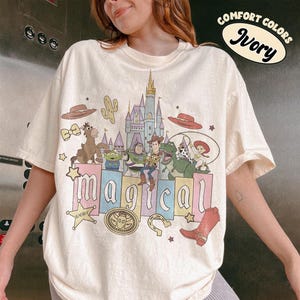 May include: Ivory-colored t-shirt featuring a colorful graphic of characters and elements from a popular animated movie. The word "magical" is displayed in large, colorful letters. The shirt also has the words "Comfort Colors" and "Ivory" printed on it.
