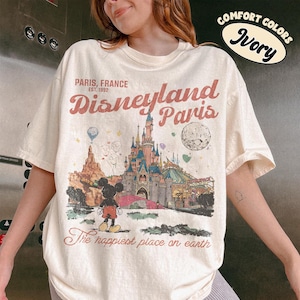 May include: Ivory-colored t-shirt featuring a Disneyland Paris graphic. The design includes a castle, Mickey Mouse, and the text "PARIS, FRANCE Disneyland Paris The happiest place on earth". The shirt is a comfort colors style.