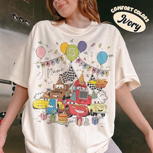 May include: Ivory-colored t-shirt with a colorful graphic featuring cartoon cars, balloons, and the text "HAPPY BIRTHDAY" and "95". The design includes a birthday cake and gift boxes, perfect for a fifth birthday celebration.