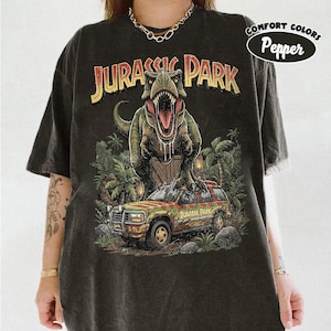 May include: Dark gray t-shirt featuring a graphic from the movie Jurassic Park. The design includes a T-Rex, a Jurassic Park vehicle, and the text "JURASSIC PARK". The shirt has a distressed look and the words "COMFORT COLORS Pepper" are printed on the upper right.