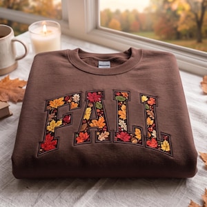 Puede incluir: Una sudadera marrón de cuello redondo con la palabra FALL en letras grandes de bloque. El interior de cada letra está relleno con un patrón detallado de coloridas hojas de otoño en tonos rojo, naranja, amarillo y marrón sobre un fondo oscuro.