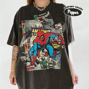 May include: Dark gray t-shirt featuring a colorful comic book design with the Amazing Spider-Man. The graphic includes the text "COMICS GROUP", "AMAZING SPIDER-MAN", and "ZING MAN". The shirt also has the text "COMFORT COLORS Pepper".