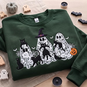 Puede incluir: Una sudadera de cuello redondo verde oscuro con un diseño bordado de tres fantasmas con patrón de encaje blanco y varios gatos negros. Un fantasma lleva un sombrero de bruja morado. El diseño incluye murciélagos blancos, telarañas y una calabaza naranja.