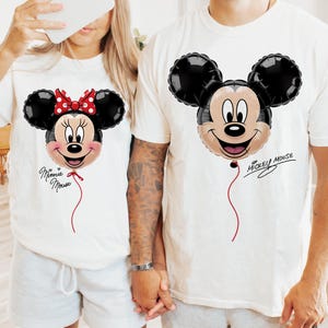 K&ouml;nnte beinhalten: Wei&szlig;e T-Shirts mit Mickey- und Minnie-Mouse-Ballon-Designs. Das Minnie-Mouse-Shirt hat eine rote Schleife und den Text "Minnie Mouse". Das Mickey-Mouse-Shirt hat den Text "Mickey Mouse". Beide Shirts haben eine rote Schnur am Ballon.