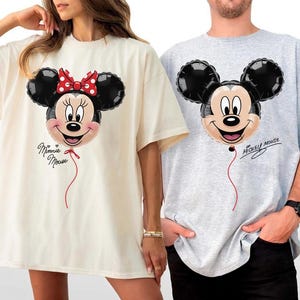 K&ouml;nnte beinhalten: Zwei &uuml;bergro&szlig;e T-Shirts, eines in Creme und eines in Grau, jeweils mit einem Cartoon-Ballon-Design. Das cremefarbene Shirt hat einen Minnie-Mouse-Ballon mit roter Schleife und dem Schriftzug "Minnie Mouse". Das graue Shirt hat einen Mickey-Mouse-Ballon.