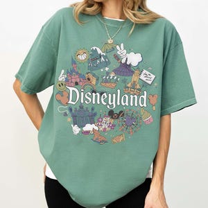 May include: A green t-shirt featuring a Disneyland graphic. The graphic includes various cartoon illustrations of Disneyland attractions, such as a castle, a Ferris wheel, and a smiling sun. The word "Disneyland" is written in a whimsical font.