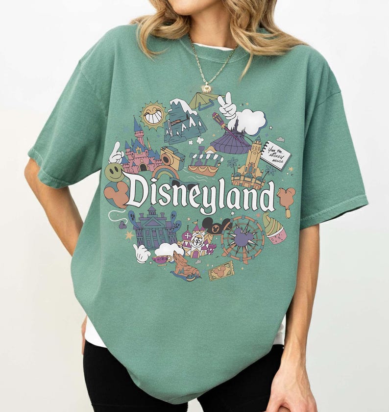 May include: A green t-shirt featuring a Disneyland graphic. The graphic includes various cartoon illustrations of Disneyland attractions, such as a castle, a Ferris wheel, and a smiling sun. The word "Disneyland" is written in a whimsical font.