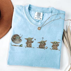 Puede incluir: Camiseta Comfort Colors azul claro con un gr&aacute;fico de un personaje en varias poses. El personaje lleva una t&uacute;nica marr&oacute;n, con una rana y una taza peque&ntilde;a. Tambi&eacute;n se ve un collar plateado con un colgante.