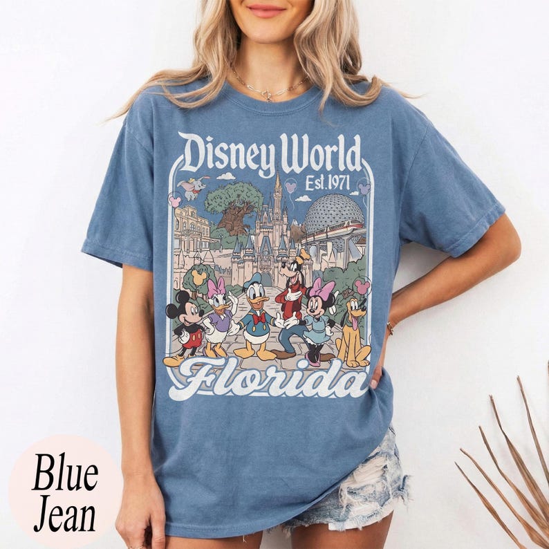 May include: Blue jean colored t-shirt featuring a Disney World graphic with Mickey Mouse, Minnie Mouse, Donald Duck, Goofy, and other characters. The text "Disney World Est. 1971 Florida" is printed on the front.