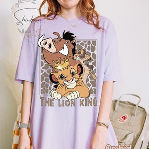 May include: A light purple t-shirt featuring a graphic of characters from The Lion King, including Simba, Pumbaa, and Timon. The words "THE LION KING" are printed below the characters in a brown font. The shirt is a relaxed fit.