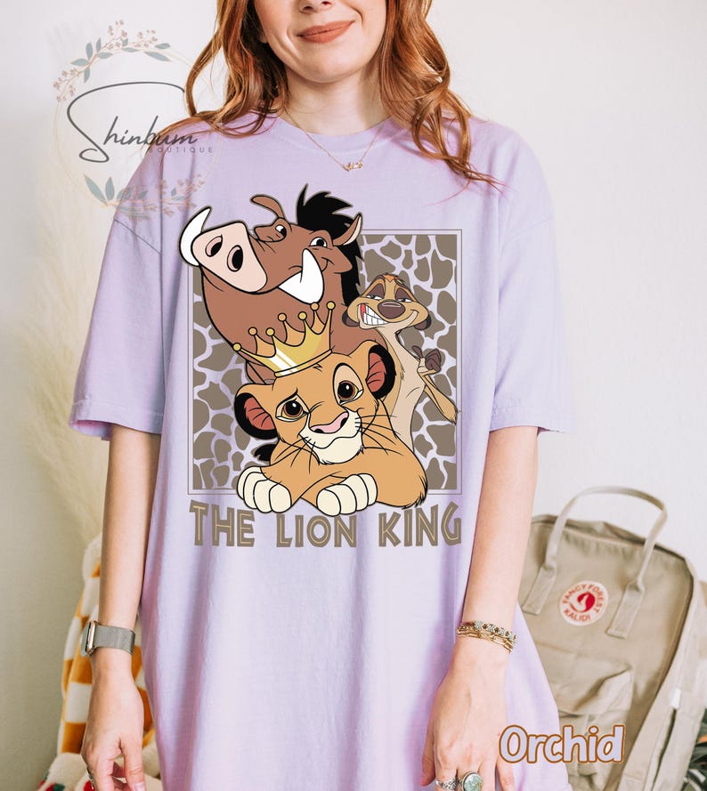 May include: A light purple t-shirt featuring a graphic of characters from The Lion King, including Simba, Pumbaa, and Timon. The words "THE LION KING" are printed below the characters in a brown font. The shirt is a relaxed fit.