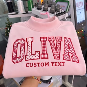 May include: A light pink mock neck sweatshirt featuring the embroidered name OLIVA in large block letters. Each letter displays a unique red and white pattern, including floral, stars, gingham, hearts, and chevron. The text CUSTOM TEXT is below.