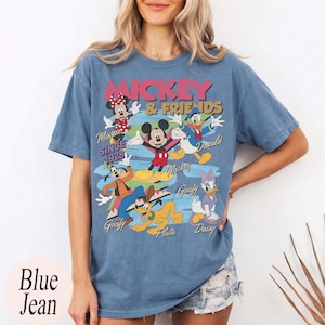 Peut inclure: T-shirt bleu jean avec un motif de Mickey Mouse et ses amis. Le motif comprend Minnie Mouse, Donald Duck, Dingo, Pluto et Daisy Duck. Le texte "Mickey & Friends" et "Since 1928" est &eacute;galement affich&eacute;.