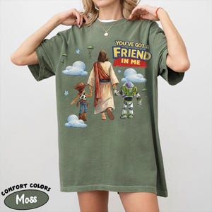 May include: Moss green t-shirt featuring the text "YOU'VE GOT A FRIEND IN ME" above an illustration of Jesus, Woody, and Buzz Lightyear. The shirt is a Comfort Colors brand.