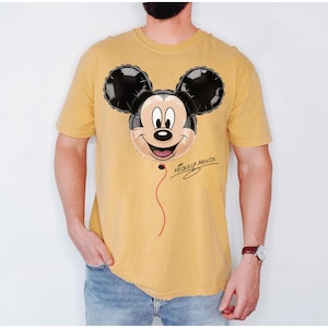 K&ouml;nnte beinhalten: Senfgelbes T-Shirt mit einem Mickey Mouse Ballon-Design. Der Ballon hat schwarze Ohren und ein l&auml;chelndes Gesicht mit einer roten Schnur. Die Worte "Mickey Mouse" sind kursiv unter dem Ballon geschrieben.