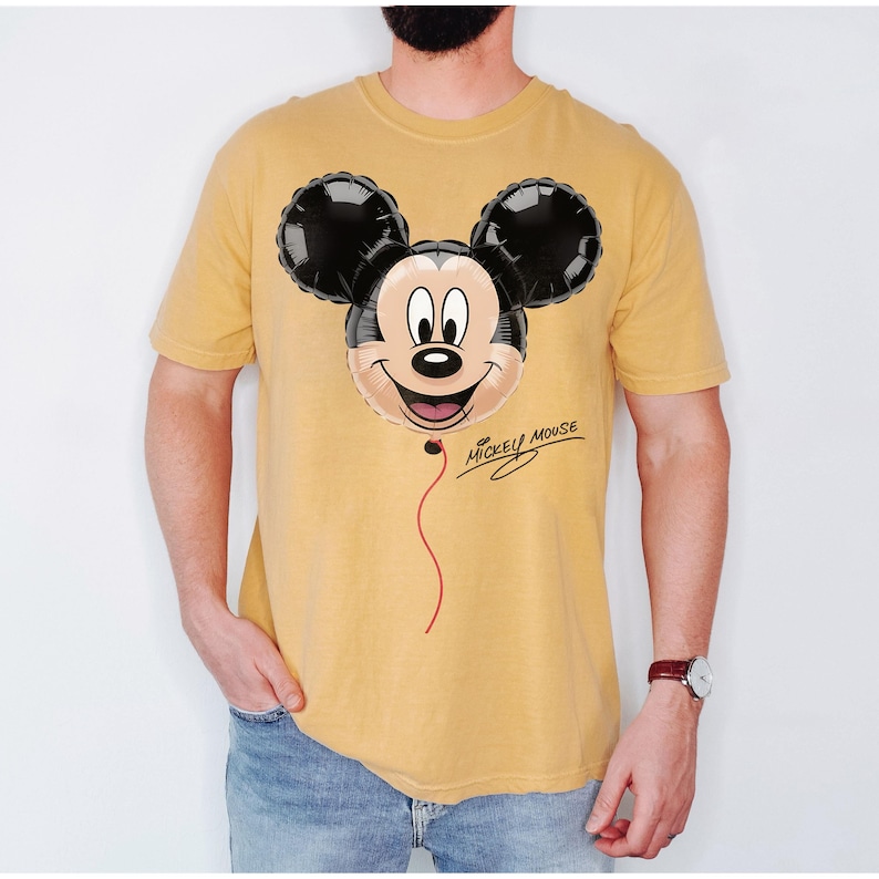 K&ouml;nnte beinhalten: Senfgelbes T-Shirt mit einem Mickey Mouse Ballon-Design. Der Ballon hat schwarze Ohren und ein l&auml;chelndes Gesicht mit einer roten Schnur. Die Worte "Mickey Mouse" sind kursiv unter dem Ballon geschrieben.