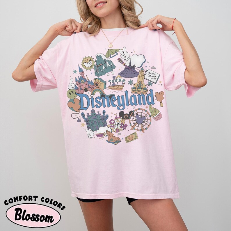 May include: A light pink t-shirt with a Disneyland graphic. The graphic features various park icons, including a castle, a Ferris wheel, and the Disneyland logo. The shirt also has the words "Comfort Colors" and "Blossom" printed on it.