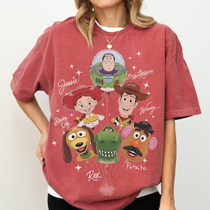 Puede incluir: Camiseta rojo-naranja con personajes de dibujos animados de la pel&iacute;cula Toy Story. El dise&ntilde;o incluye a Jessie, Buzz Lightyear, Woody, Slinky Dog, Rex y Mr. Potato. Los nombres de los personajes est&aacute;n escritos en una fuente l&uacute;dica.