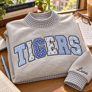 May include: A heather gray sweatshirt with a mock neck and cuffs featuring a gray and white gingham pattern. The front displays the word TIGERS in large block letters with varied blue and white floral, striped, and polka dot patterns. The name Luna is embroidered in dark blue script on the right sleeve.